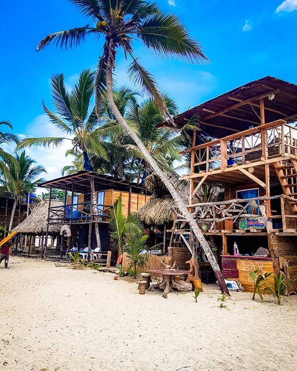 Rustic wooden beach huts on a sandy beach surrounded by palm trees under a bright blue sky. The huts have thatched roofs, open-air design, and a mix of seating areas, with a relaxed, tropical vibe.