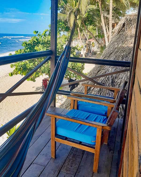 A cozy balcony overlooking the beach, featuring a hammock and a wooden chair with bright blue cushions. The view includes lush greenery, palm trees, and the ocean waves crashing onto the sandy shore.
