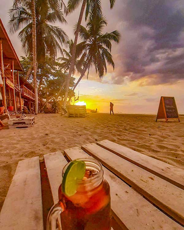 A sunset scene on the beach with palm trees and a wooden lounge area, featuring a drink with a lime wedge in the foreground. The sky is a mix of golden and dusky hues as the sun sets, with a person walking along the shore and beach huts visible in the background.